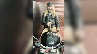 Longhairdude masturbating and shooting sitting astride Motorcycle in leather jacket, motocross footwear, hat